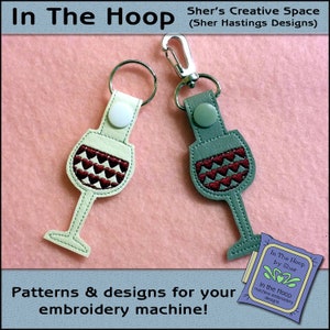 May include: Two wine glass keychains with red and white heart embroidery. The keychains are made of leather and have a silver metal ring. The keychains are on a pink background. The text "In The Hoop by Sher" is on a purple tag.