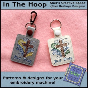 May include: Two gray and white keychains with a cross and the words "Just Pray" embroidered on them. The keychains are on a pink fabric background. The text "In The Hoop by Sher" is visible in the lower right corner of the image.