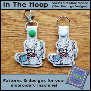 Peut inclure: Deux porte-clés blancs avec des images brodées d'un chef tenant un bol et une cuillère. Les porte-clés sont attachés à des anneaux de clés argentés. Le texte "In The Hoop by Sher" est visible sur une petite étiquette.