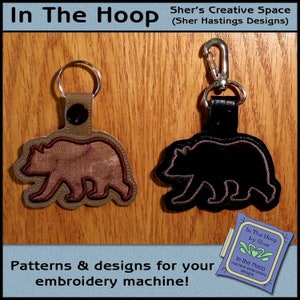 May include: Two bear-shaped keychains. One is brown with a tan outline and a silver key ring. The other is black with a silver clasp. The text "In The Hoop" and "Patterns & designs for your embroidery machine!" are also visible.