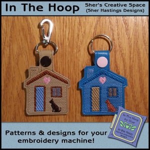 May include: Two house-shaped keychains. One is tan with a brown roof, blue door, and black cat. The other is blue with a brown door and orange cat. Both have a pink heart and window. The keychains are attached to a metal clasp and ring.