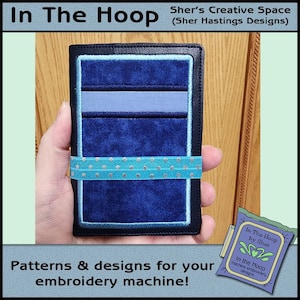 May include: A blue and gray embroidered wallet with a turquoise ribbon and white polka dots. The wallet is held in a hand. The text "In The Hoop by Sher" is visible in the lower right corner of the image.