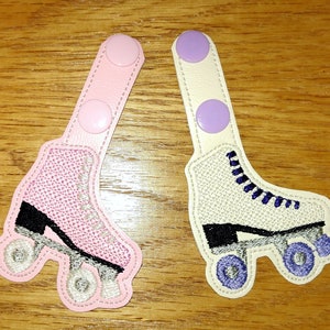 K&ouml;nnte beinhalten: Zwei Paare von Rollschuh-f&ouml;rmigen Stoff-Patches mit Klettverschl&uuml;ssen. Ein Paar ist rosa mit schwarzen R&auml;dern und einem schwarzen Rollschuh. Das andere Paar ist wei&szlig; mit lila R&auml;dern und einem schwarzen Rollschuh.