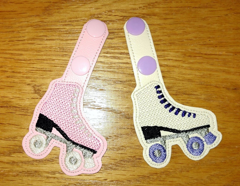 K&ouml;nnte beinhalten: Zwei Paare von Rollschuh-f&ouml;rmigen Stoff-Patches mit Klettverschl&uuml;ssen. Ein Paar ist rosa mit schwarzen R&auml;dern und einem schwarzen Rollschuh. Das andere Paar ist wei&szlig; mit lila R&auml;dern und einem schwarzen Rollschuh.