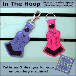 May include: Two keychains in the shape of lingerie, one purple and one pink, with black lace trim. The keychains are attached to silver metal clasps and rings. The image includes text that reads "In The Hoop" and "Patterns & designs for your embroidery machine!"