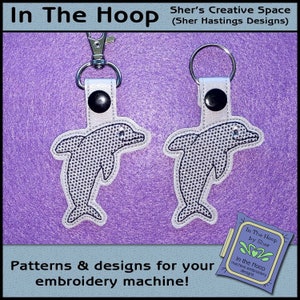 Puede incluir: Dos llaveros de delfín blanco con contornos negros y un patrón de cuadrícula blanco y negro. Los llaveros están unidos a llaveros plateados. La imagen incluye el texto "In The Hoop by Sher" y "In the Hoop machine embroidery designs".