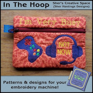 May include: A small orange zippered pouch with a red zipper. The pouch has an embroidered design of a video game controller and headphones with the text "I'm Very Busy" and "Not Now".