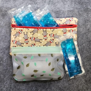 K&ouml;nnte beinhalten: Eine beige Rei&szlig;verschlusstasche mit rotem Rei&szlig;verschluss, gef&uuml;llt mit blauen K&uuml;hlpacks. Die Tasche hat ein Muster aus grauen Sockenaffen mit roten und wei&szlig;en H&uuml;ten. Vorne ist eine transparente Plastikt&uuml;te mit gr&uuml;nen Bl&auml;ttern.