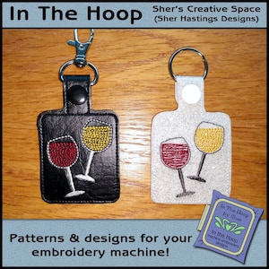 May include: Two wine glass keychains, one black and one white, with red and yellow wine glasses embroidered on them. The keychains are attached to key rings. The text "In The Hoop by Sher" is visible in the lower right corner of the image.