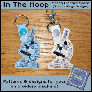 May include: Two microscope-shaped keychains. One is light blue with a teal snap, the other is off-white with a gray snap. Both have silver key rings and are embroidered with black and gray details. The image includes text: "In The Hoop" and "Patterns & designs for your embroidery machine!"