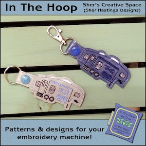 May include: Two embroidered camper van keychains, one in blue and one in white, with a blue and white button on each. The keychains are on a wooden surface. The text "In The Hoop by Sher" is visible in the lower right corner of the image.