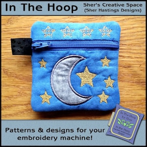 May include: A blue zippered pouch with a crescent moon and stars embroidered on it. The pouch is on a wooden surface. The text "In The Hoop by Sher" is on a small tag in the lower right corner of the image.