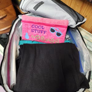 K&ouml;nnte beinhalten: Ein offener schwarzer Rucksack zeigt eine graue Rei&szlig;verschlusstasche, eine pinkfarbene Rei&szlig;verschlusstasche mit dem Text "COOL STUFF" und gefaltete schwarze Kleidung. Die pinkfarbene Tasche hat ein Thermometer- und Sonnenbrillen-Design.