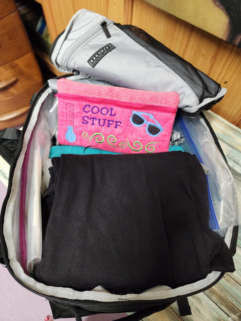 K&ouml;nnte beinhalten: Ein offener schwarzer Rucksack zeigt eine graue Rei&szlig;verschlusstasche, eine pinkfarbene Rei&szlig;verschlusstasche mit dem Text "COOL STUFF" und gefaltete schwarze Kleidung. Die pinkfarbene Tasche hat ein Thermometer- und Sonnenbrillen-Design.