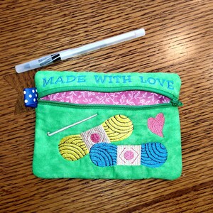 May include: Green zippered pouch with the text "Made with Love" embroidered on the top. The pouch has a pink and white floral lining and features embroidered images of yarn, a crochet hook, and a heart.