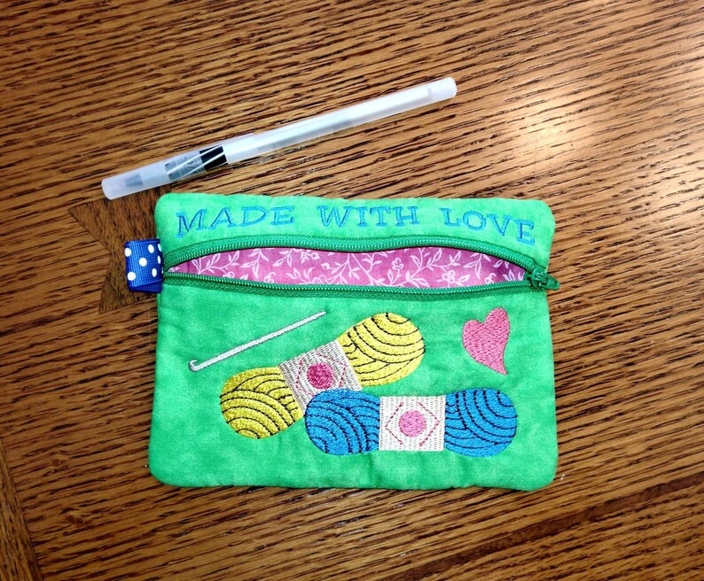 May include: Green zippered pouch with the text "Made with Love" embroidered on the top. The pouch has a pink and white floral lining and features embroidered images of yarn, a crochet hook, and a heart.