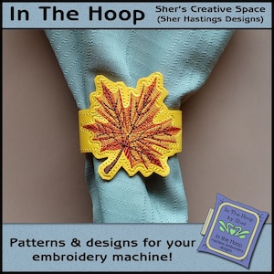 May include: A yellow embroidered maple leaf napkin ring on a light blue napkin. The text "In The Hoop by Sher" is visible in the lower right corner of the image.