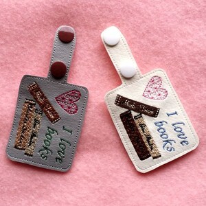May include: Two gray and white fabric keychains with embroidered images of stacked books and hearts. The text "I love books" is embroidered on the white keychain.