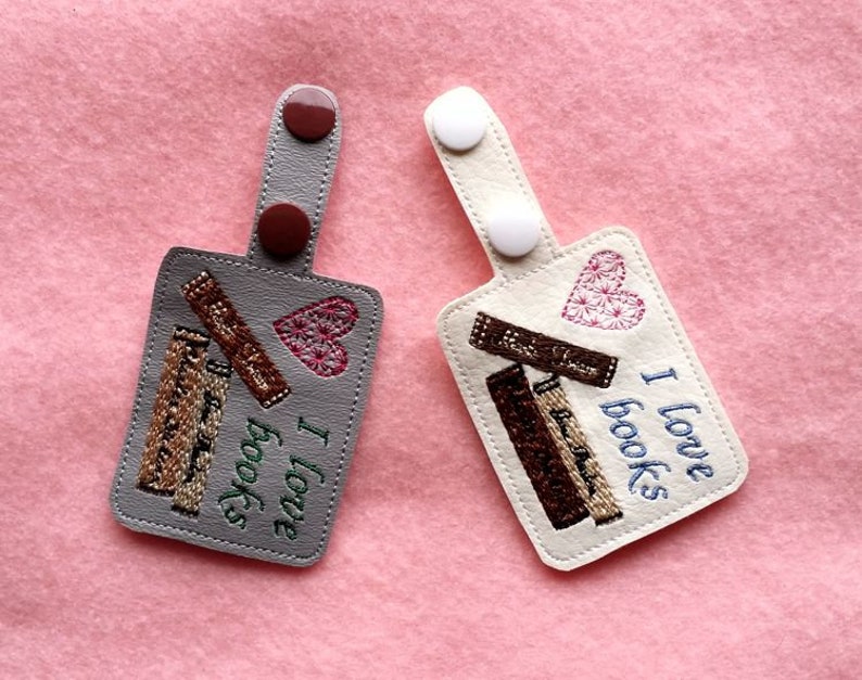 May include: Two gray and white fabric keychains with embroidered images of stacked books and hearts. The text "I love books" is embroidered on the white keychain.