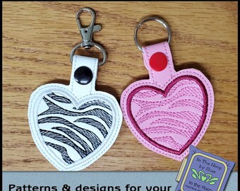Puede incluir: Dos llaveros en forma de corazón con un diseño de estampado de cebra. El llavero de la izquierda es blanco con rayas negras y el llavero de la derecha es rosa con rayas rojas. Ambos llaveros tienen un anillo de llave plateado y un botón negro.