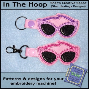 May include: Two pink and purple embroidered keychains with sunglasses designs. The keychains are on a light blue background. The text "In The Hoop by Sher" is visible in the lower right corner of the image.