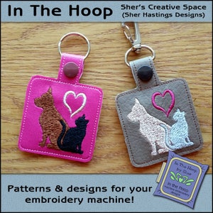 May include: Two keychains with embroidered designs of a dog and cat with a heart. The keychain on the left is pink with brown and black embroidery. The keychain on the right is gray with pink and blue embroidery. The text "In The Hoop by Sher" is visible on a small tag.