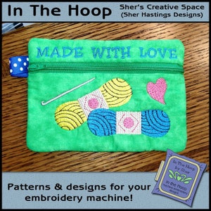 May include: Green zippered pouch with the text "MADE WITH LOVE" embroidered on the front. The pouch features an embroidered design of two balls of yarn, a needle, and a heart.