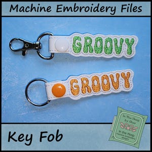 May include: Two white key fobs with the word "GROOVY" embroidered in green and orange thread. Each fob has a metal ring or clasp and a colored button. The text "Machine Embroidery Files" and "Key Fob" are also visible.