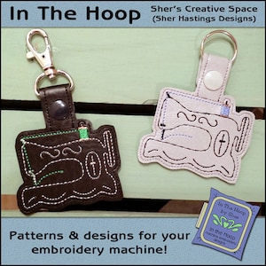 May include: Two black and white sewing machine keychains with a green and blue thread detail. The keychains are on a wooden surface. The text "In The Hoop by Sher" is on a purple and green label.