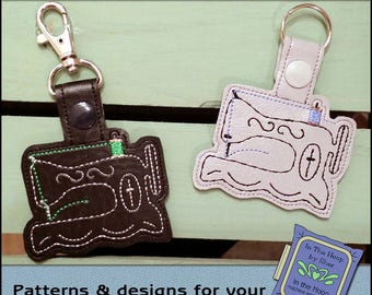May include: Two black and white sewing machine keychains with a green and blue thread detail. The keychains are on a wooden surface. The text "In The Hoop by Sher" is on a purple and green label.