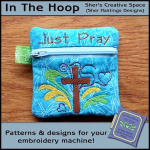 May include: A blue zippered pouch with a cross and hearts embroidered on it. The pouch has the words "Just Pray" embroidered on the top. The pouch is on a wooden surface.