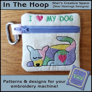 May include: A small, white zippered pouch with a colorful embroidered dog design. The pouch has a pink zipper pull and a silver carabiner clip. The text "I ♥ MY DOG" is embroidered on the top of the pouch.