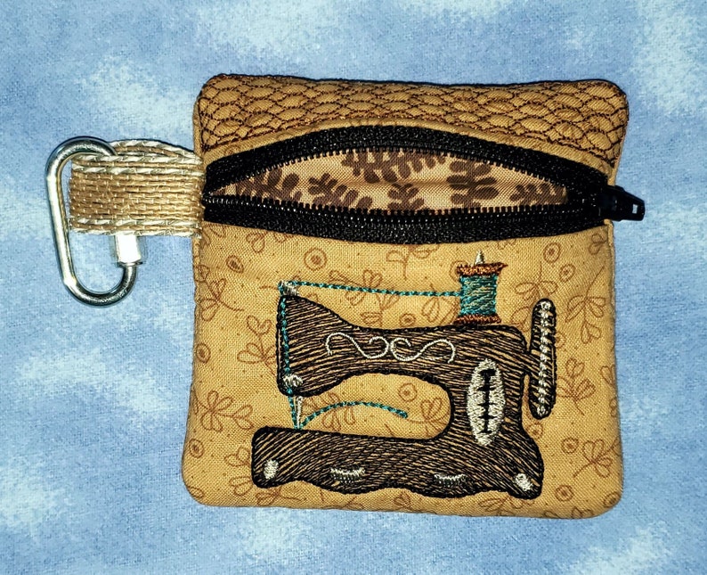 May include: A small brown zippered pouch with a brown and black sewing machine design. The pouch has a brown and black zipper and a silver carabiner clip.