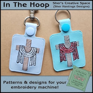 May include: Two keychains with embroidered crosses and fabric draped over them. The keychains are on a brown background. The text "In The Hoop by Sher" is visible in the lower right corner of the image.