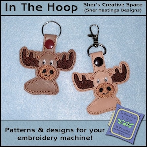 May include: Two brown moose keychains with embroidered details. The keychains are attached to silver key rings. The keychains are on a light blue background. The text "In The Hoop by Sher" is visible in the lower right corner of the image.