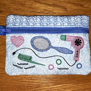 May include: A blue zippered pouch with a white floral pattern. The pouch has embroidered images of a hairdryer, a comb, a hairbrush, a mirror, and a heart.