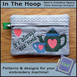 Puede incluir: Una bolsa gris con cremallera y diseño acolchado. La bolsa presenta un diseño bordado de una tetera y una taza de té con corazones y el texto "It's always tea time".