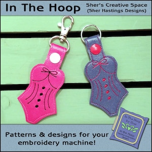May include: Two embroidered keychains in the shape of a corset. The keychain on the left is pink with black stitching and the keychain on the right is purple with red stitching. The keychains are on a wooden background with the text "In The Hoop by Sher" and "In the Hoop machine embroidery designs".