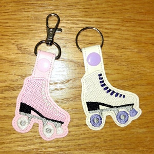 K&ouml;nnte beinhalten: Zwei gestickte Rollschuh-Schl&uuml;sselanh&auml;nger. Einer ist pink mit schwarzen R&auml;dern und der andere ist wei&szlig; mit lila Schn&uuml;rsenkeln und schwarzen R&auml;dern.