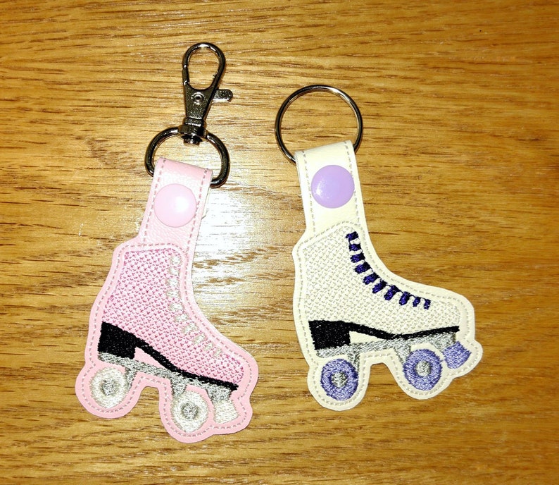 K&ouml;nnte beinhalten: Zwei gestickte Rollschuh-Schl&uuml;sselanh&auml;nger. Einer ist pink mit schwarzen R&auml;dern und der andere ist wei&szlig; mit lila Schn&uuml;rsenkeln und schwarzen R&auml;dern.
