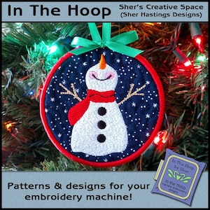 May include: A round, blue and white embroidered ornament featuring a snowman with a red scarf and a starry background. The ornament is hanging on a Christmas tree with lights. The text "In The Hoop by Sher" is visible in the lower right corner of the image.