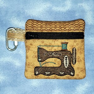 May include: A small brown zippered pouch with a brown and black embroidered sewing machine design. The pouch has a brown and black zipper and a brown loop.