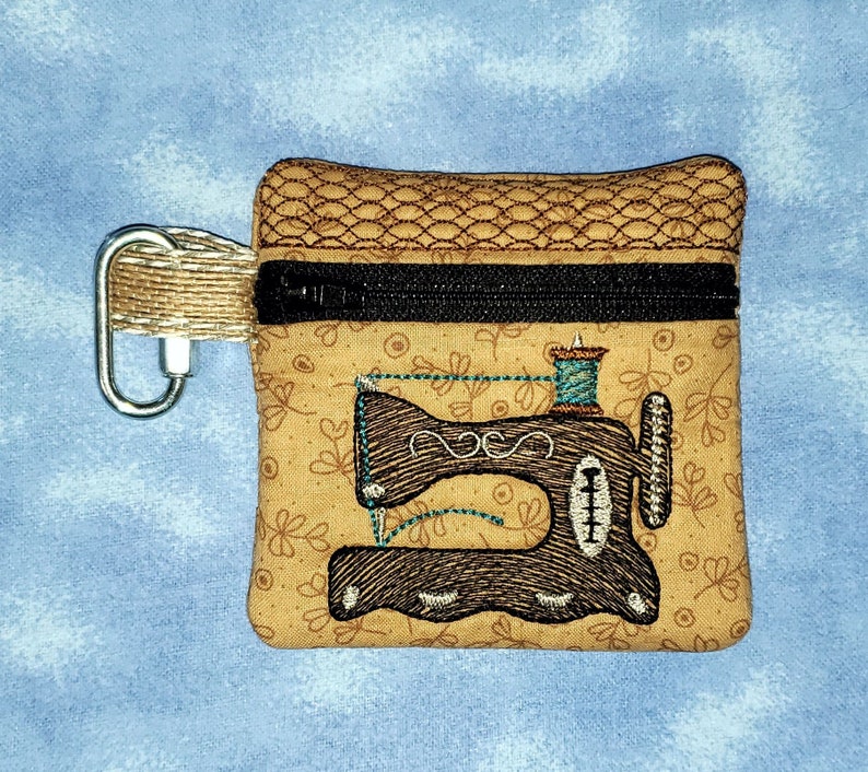 May include: A small brown zippered pouch with a brown and black embroidered sewing machine design. The pouch has a brown and black zipper and a brown loop.