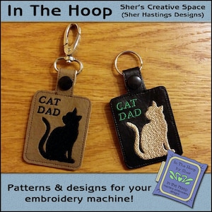 Peut inclure: Deux porte-clés avec des motifs de chat brodés. Le porte-clés de gauche a un fond brun avec un chat noir et le texte "CAT DAD". Le porte-clés de droite a un fond noir avec un chat beige et le texte "CAT DAD" en vert. Les deux porte-clés ont un anneau en métal argenté.