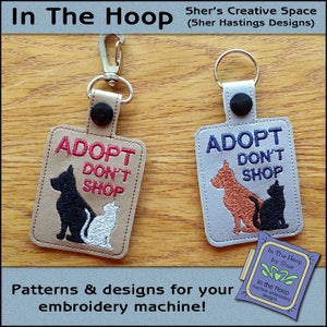 May include: Two keychains with embroidered designs featuring a dog and cat with the text "Adopt Don't Shop". The keychains are beige and gray with black and white embroidery.