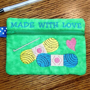 May include: Green zippered pouch with the text "MADE WITH LOVE" embroidered on the top. The pouch features an embroidered design of two balls of yarn, a crochet hook, and a heart.