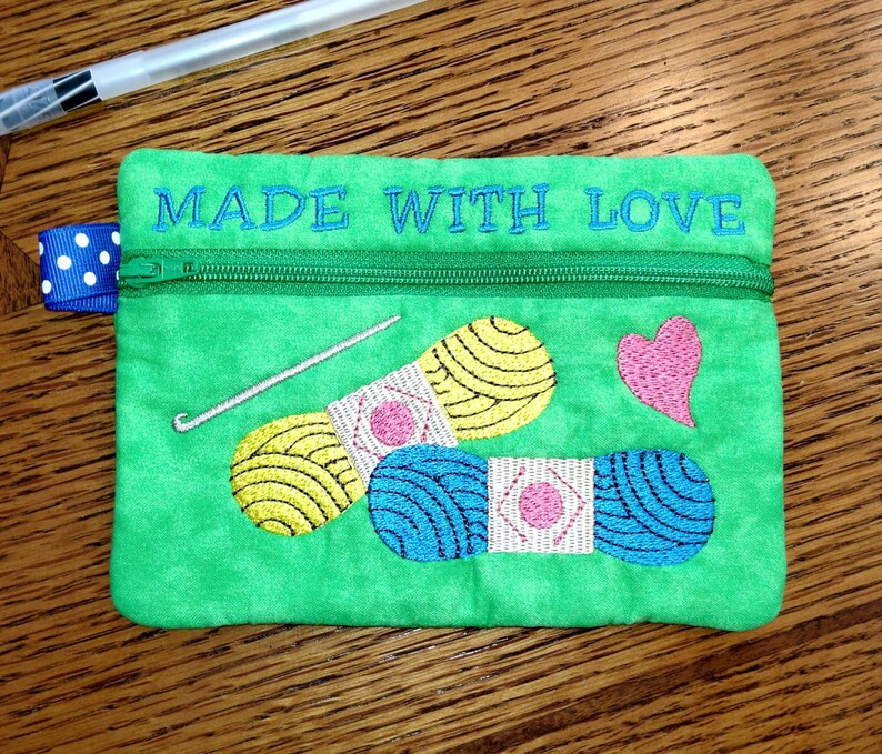 May include: Green zippered pouch with the text "MADE WITH LOVE" embroidered on the top. The pouch features an embroidered design of two balls of yarn, a crochet hook, and a heart.