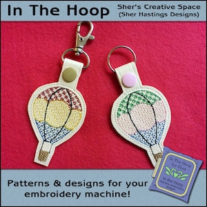May include: Two hot air balloon keychains with embroidery designs. The keychains are on a red felt background. The text "In The Hoop by Sher" is visible in the lower right corner of the image.