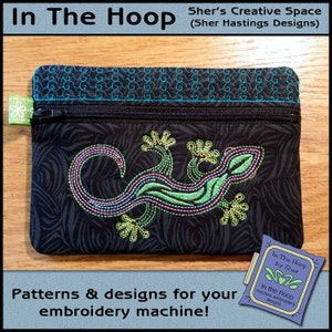 Könnte beinhalten: Eine schwarze Reißverschlusstasche mit einem gestickten grünen und gelben Eidechsen-Design. Die Tasche liegt auf einer Holzoberfläche. Der Text "In The Hoop by Sher" befindet sich auf einem blau-weißen Etikett.
