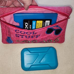 K&ouml;nnte beinhalten: Rosa isolierte K&uuml;hltasche mit dem gestickten Text "COOL STUFF" und Sonnenbrille. Die Tasche enth&auml;lt einen blauen K&uuml;hlakku. Ein zweiter blauer K&uuml;hlakku befindet sich unter der Tasche. Die Tasche ist ca. 10,8 x 21 cm gro&szlig;.